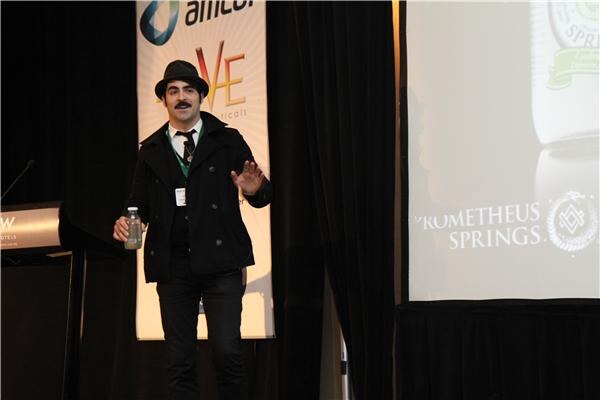 A mustachioed man in a black hat and coat holds a bottle and gestures on stage. A screen beside him shows the Prometheus Springs name and logo.