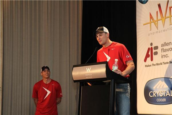 A man in a red Nike shirt and cap speaks at a W Hotels podium while holding a water bottle. Another man in red smiles behind him, with NVE, Allen Flavors and Crystal Cooler banners.