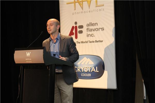 A bald man in a gray blazer and blue shirt speaks at a W Hotels podium beside a banner with Allen Flavors Inc. and Crystal Cooler logos.