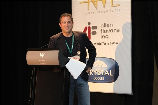 A man in a dark jacket and green lanyard stands at a W Hotels podium holding papers and a water bottle, in front of a banner for Allen Flavors Inc. and Crystal Cooler.