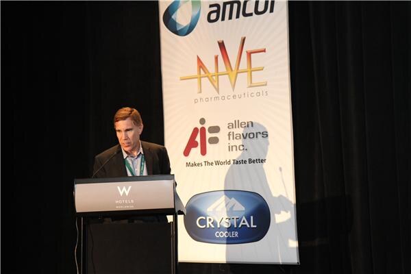 A man in a dark jacket speaks at a W Hotels podium in front of a banner showing Amcor, NVE Pharmaceuticals, Allen Flavors Inc. and Crystal Cooler logos.