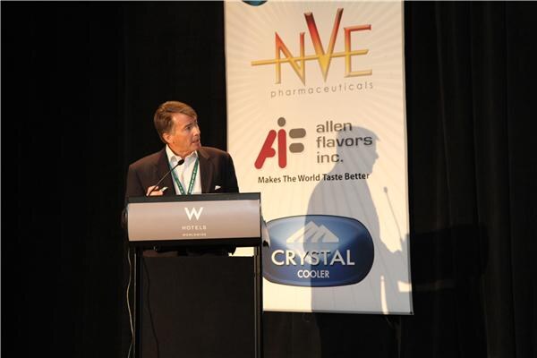A man in a dark blazer speaks at a podium. A banner behind him shows NVE Pharmaceuticals, Allen Flavors Inc. and Crystal Cooler logos.