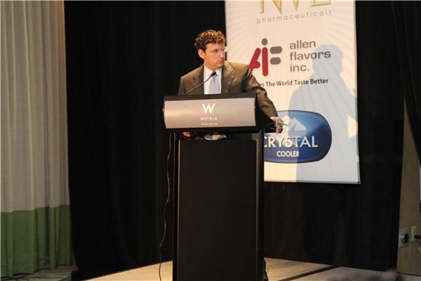 A man with curly hair, wearing a suit and glasses, stands at a W Hotels podium and looks to the side, beside a banner for Allen Flavors Inc. and Crystal Cooler.