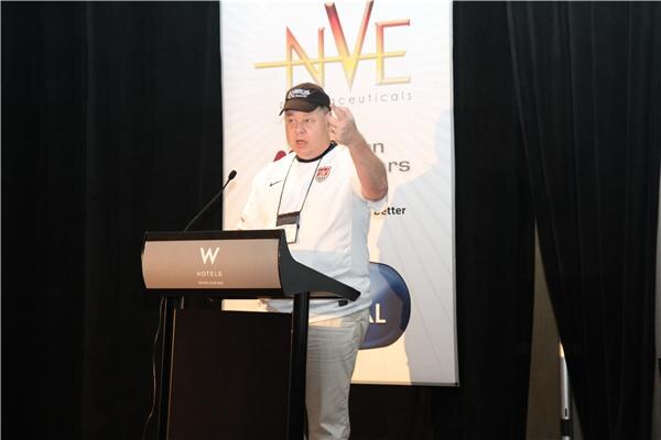 A man in a cap and white soccer jersey gestures while speaking at a W Hotels podium in front of a banner with the NVE Pharmaceuticals logo.