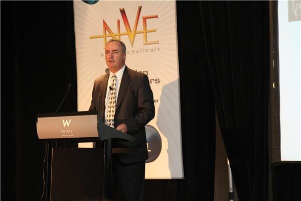 A man in a dark suit and patterned tie speaks at a W Hotels podium in front of a banner with the NVE Pharmaceuticals logo.
