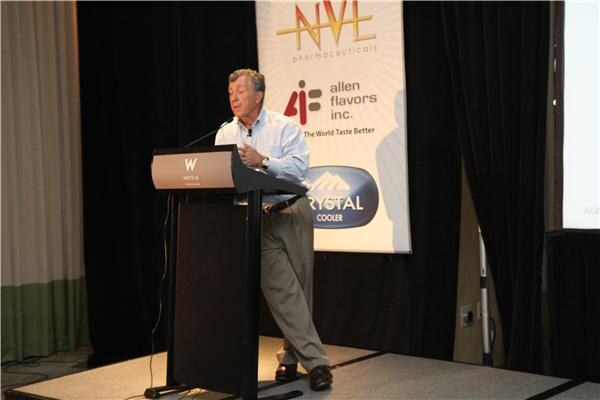 A man in a light blue shirt and khaki pants leans on a W Hotels podium on a stage, in front of a banner with NVE Pharmaceuticals, Allen Flavors Inc. and Crystal Cooler logos.