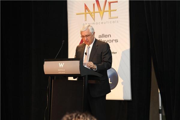 A gray-haired man in glasses and a dark suit looks down at a W Hotels podium in front of a banner showing NVE Pharmaceuticals and Allen Flavors logos.
