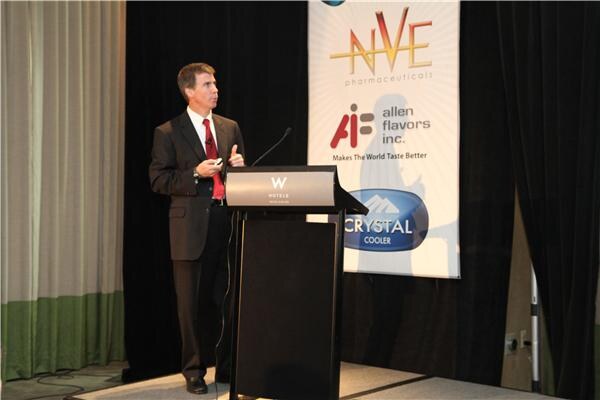 A man in a dark suit and red tie holds a clicker and gestures beside a W Hotels podium, in front of a banner for NVE Pharmaceuticals, Allen Flavors Inc. and Crystal Cooler.