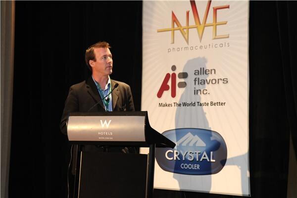 A man in a dark jacket stands at a W Hotels podium and looks to his left, in front of a banner for NVE Pharmaceuticals, Allen Flavors Inc. and Crystal Cooler.