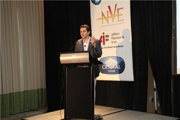 A man in a dark blazer gestures while speaking at a W Hotels podium, in front of a banner for NVE Pharmaceuticals, Allen Flavors Inc. and Crystal Cooler.