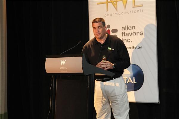 A man in a black polo and white pants holds a small bottle while speaking at a W Hotels podium in front of a banner with the Allen Flavors Inc. logo.