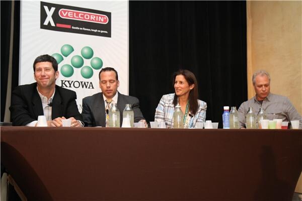 Two men and a woman sit at a brown draped table with water bottles and cups, with a fourth man at right. Velcorin and Kyowa signage is behind them.