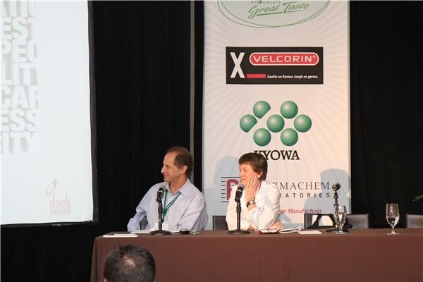 A man and a woman sit at a paneled table on stage. A banner behind them shows Velcorin and Kyowa logos, and a projection screen is at left. The back of an audience member's head is in the foreground.