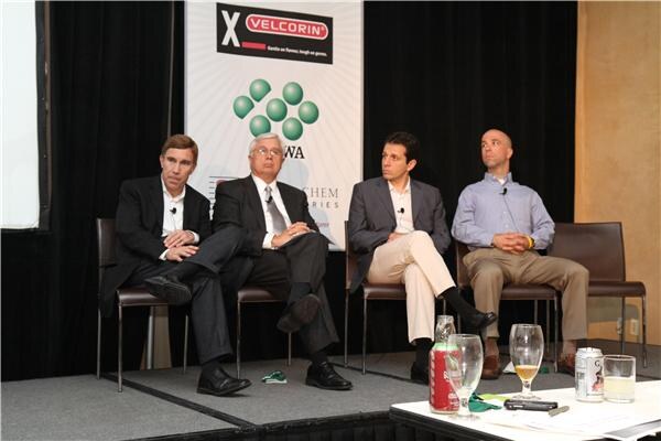 Four men in business attire sit on chairs on a stage with a Velcorin banner behind them. A low table in front holds glasses, a can and a bottle.