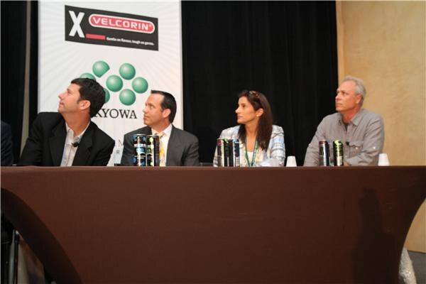 Three men and a woman sit behind a draped table holding energy drink cans, looking up and to the left. A Velcorin banner is behind them.