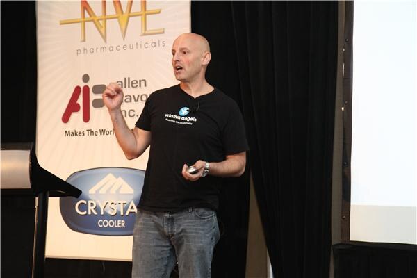 A bald man in a black Vitamin Angels t-shirt and jeans gestures while speaking, holding a clicker, in front of a banner for NVE Pharmaceuticals, Allen Flavors Inc. and Crystal Cooler.