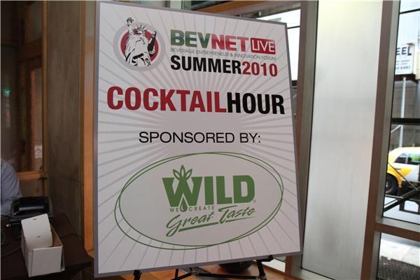 A sign on an easel reads BevNET Live Summer 2010 Cocktail Hour, sponsored by WILD, We Create Great Taste. A yellow taxi is visible through the window.