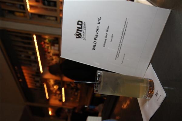 A drink in a tall glass with a straw sits on a W Hotels napkin beside a printed WILD Flavors, Inc. card titled Whiskey Sour Mixer, with a bar in the background.