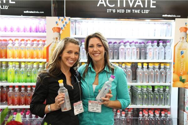 Two smiling women with show badges stand together in the Activate booth, one holding a bottle. Shelves of colorful Activate drinks are behind them.