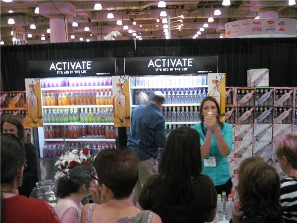 A woman in a teal top speaks to a crowd in front of the Activate booth, whose lit shelves hold rows of bottled drinks. Attendees in the foreground face the booth.