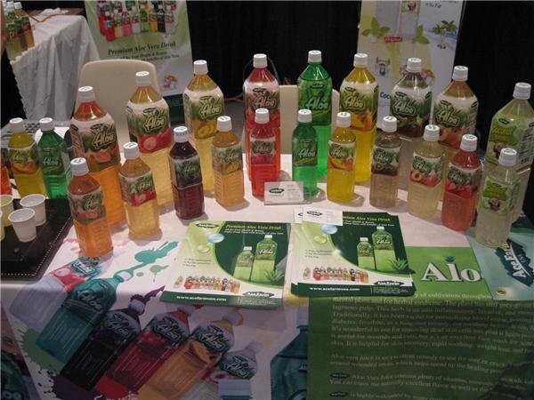 A table displays many bottles of Alo aloe vera drink in assorted flavors, with brochures in front and small sample cups at left.