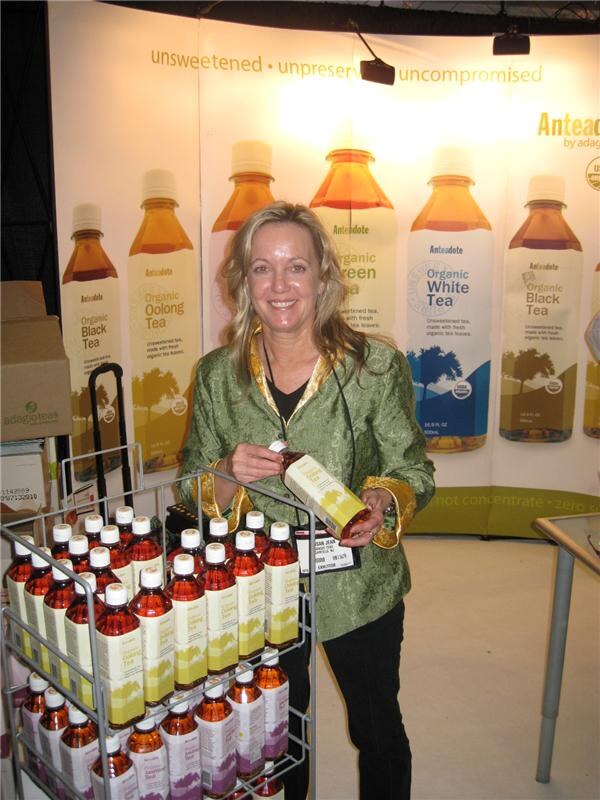 A smiling woman in a green jacket holds a boxed tea bottle beside a rack of Anteadote bottles. Banners behind her show organic black, oolong, green and white tea bottles.
