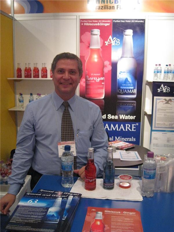 A smiling man in a blue shirt and tie stands behind a blue table holding Aquamare purified sea water bottles, with a Maryam hibiscus and ginger bottle and brochures in front of an Aquamare poster.