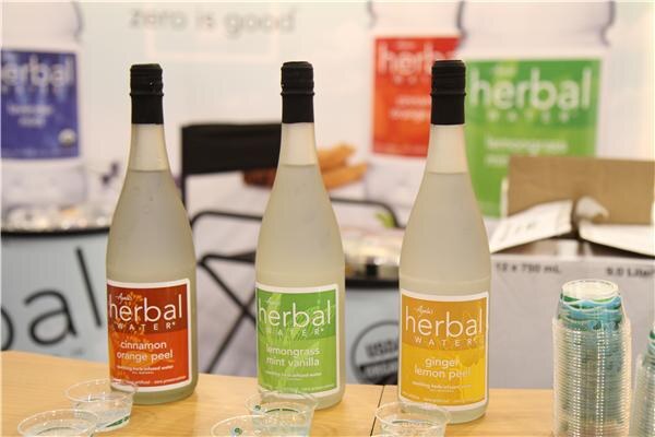 Three bottles of Ayala's Herbal Water in cinnamon orange peel, lemongrass mint vanilla and ginger lemon peel stand on a table with small sample cups and a stack of cups, in front of herbal water signage.
