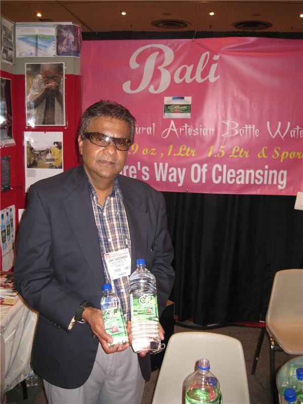 A man in dark-tinted glasses and a dark blazer holds two bottles of Bali natural artesian bottled water in front of a pink Bali banner at a trade show booth.