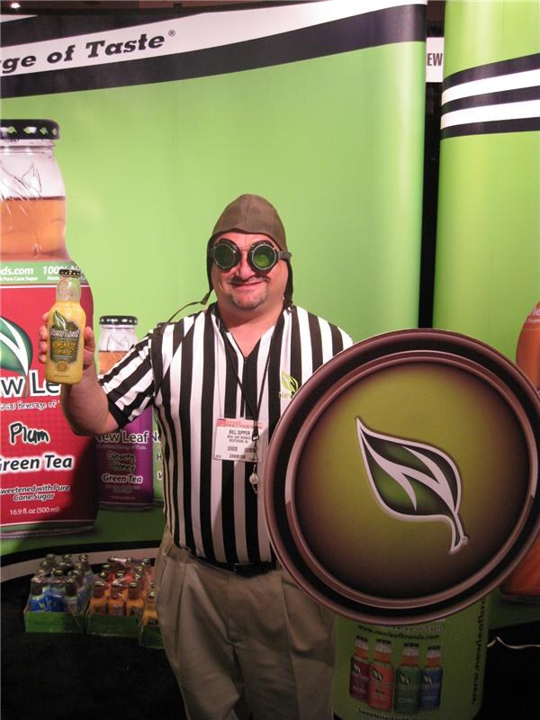 Bill Sipper of New Leaf, dressed in a referee shirt, aviator cap and goggles, holds a bottle of New Leaf tea and a large leaf-logo shield in front of a green New Leaf banner.