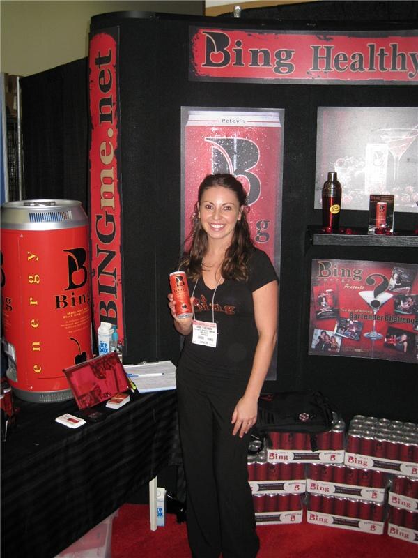 A smiling woman in a black Bing shirt holds a red Bing energy drink can in a booth with a large red can display, a BINGme.net banner, and cases of Bing cans stacked on the floor.