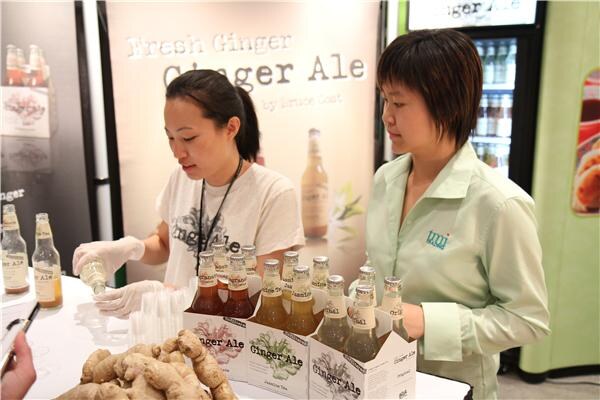 Two women work at a Bruce Cost Ginger Ale booth, one pouring samples with gloved hands, with cartons of ginger ale bottles and fresh ginger roots on the table below a Fresh Ginger Ale sign.