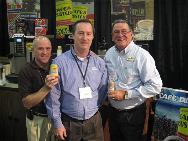 Three smiling men stand together in a Café Bustelo booth, two holding small cans of Café Bustelo coffee. Café Bustelo and Supreme by Bustelo signs and an espresso machine are behind them.