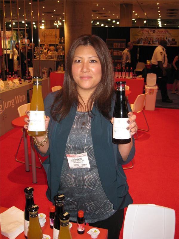 Cathlin Lee of Daesang America holds a bottle of yellow drink in one hand and a bottle of dark red drink in the other, with more bottles and sample cups on a red table in front of her.