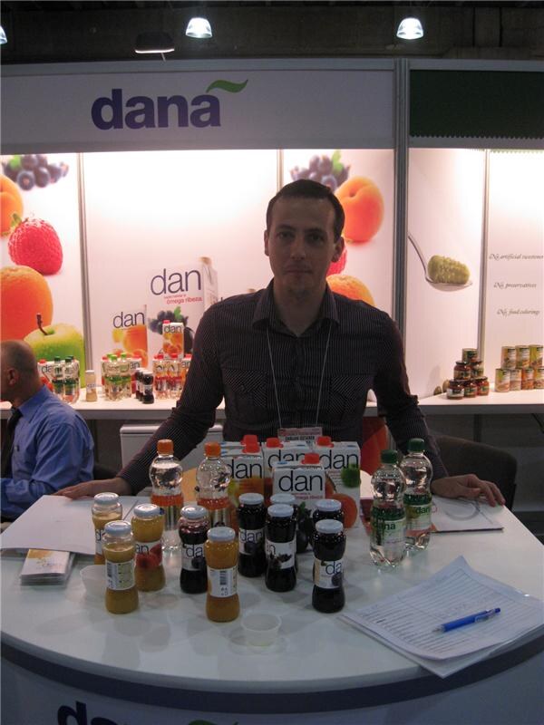 A man in a dark striped shirt sits behind a curved white counter under a dana sign, with dan juice cartons, bottles and jars of fruit products, and papers with a pen in front of him.