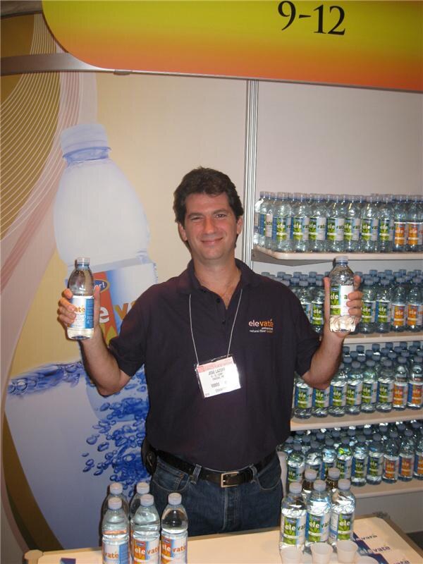 A smiling man in a dark polo with a show badge holds up two Elevate water bottles in a booth. Shelves of Elevate bottles and a large bottle graphic are behind him.