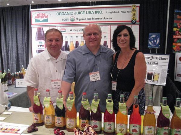 Two men and a woman stand together behind a table of Elite Naturel juice bottles with green neck labels, below an Organic Juice USA Inc. banner.