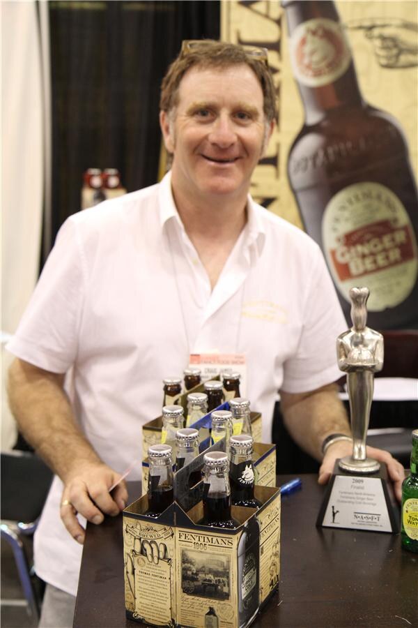 A smiling man in a white shirt stands behind a table with Fentimans six-pack bottle carriers and a silver award trophy. A large Fentimans Ginger Beer bottle graphic hangs behind him.