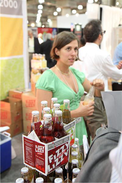 A woman in a green dress holds a bottle beside a Fizzy Lizzy four-pack carrier with more bottles in front of her. A man in a white shirt stands in the background among boxes.