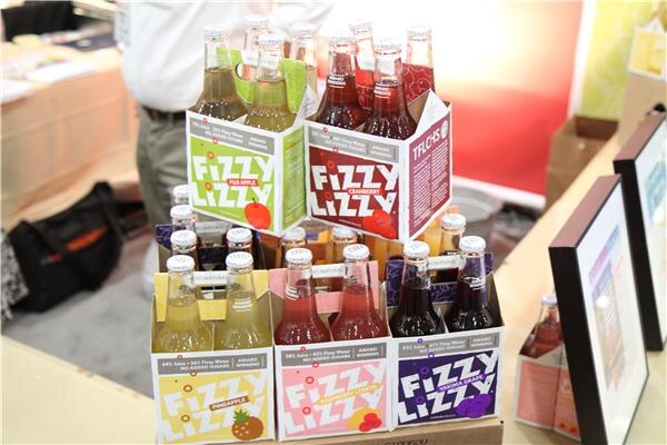Fizzy Lizzy four-pack carriers in apple, cranberry, pineapple, raspberry lime and grape flavors are stacked in a display, with framed signs at the right edge.