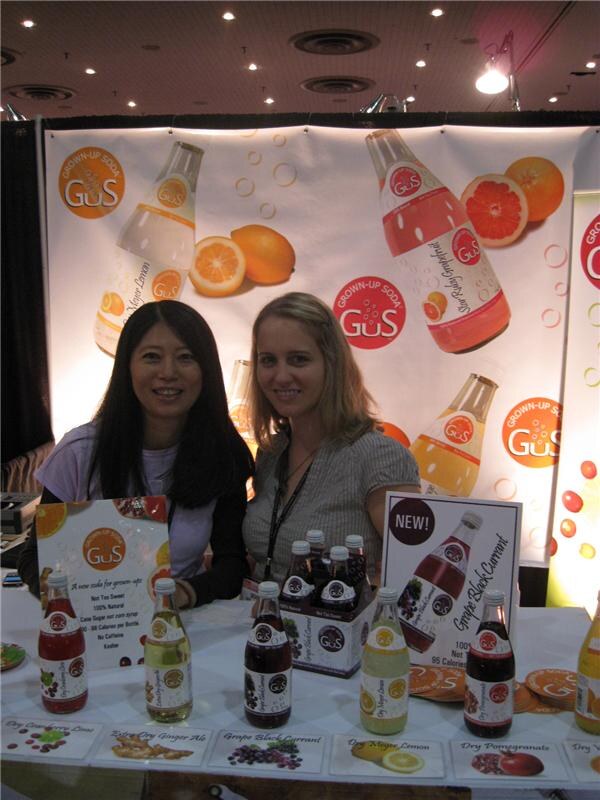 Two smiling women sit behind a table of Grown-Up Soda GuS bottles in front of a backdrop showing bottles and citrus slices. A sign promotes the new Grape Black Currant flavor.