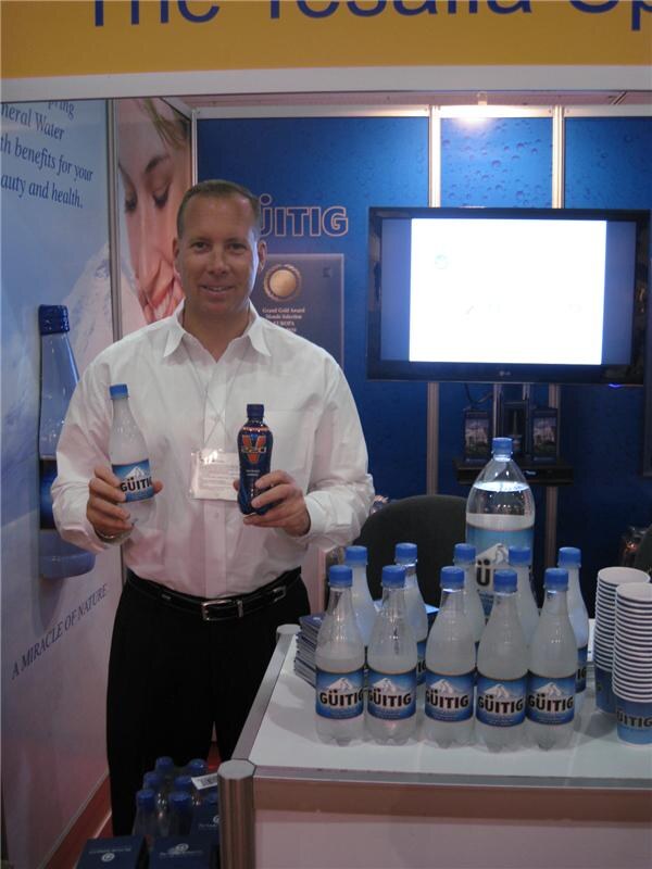 A smiling man in a white shirt holds a bottle of Güitig mineral water and a dark bottle, with Güitig bottles on a table in front of him and a blue backdrop with a monitor behind.