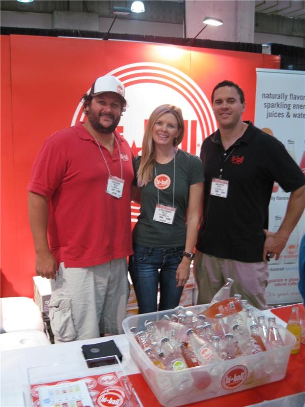 Two men and a woman wearing badges stand together at a red Hi-Ball booth. A tub of iced bottles sits on the table in front of them.