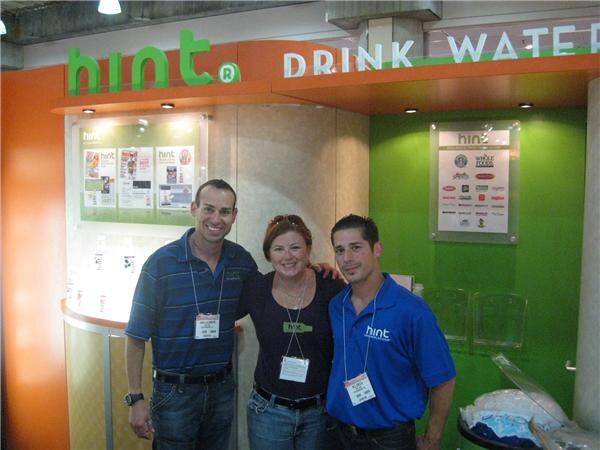 Two men and a woman wearing badges stand together in front of a Hint booth with green and orange signage reading Drink Water.