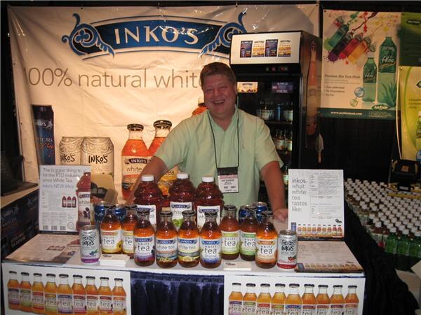A smiling man in a light green shirt stands behind a table lined with Inko's white tea bottles and cans, in front of a banner reading 100% natural white tea.