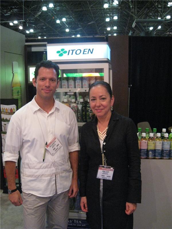 A man in a white shirt and a woman in a black jacket stand side by side in front of a lit Ito En display cooler holding bottled tea, with more bottles on a counter at right.