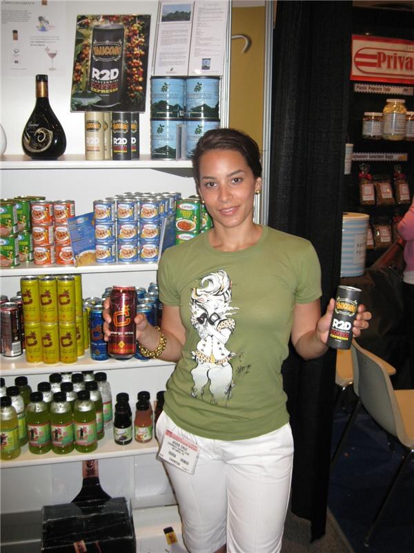 Jessie Cruz, wearing a green T-shirt and white pants, holds two energy drink cans, one marked R2D, in front of shelves of canned goods and bottled drinks.