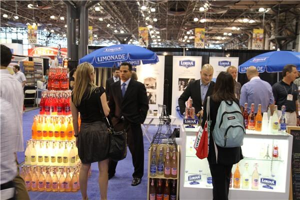 Visitors walk past a Lorina booth with blue Limonade umbrellas, tiered displays of orange, yellow and red bottles, and a lit counter of bottles where a man in a dark suit stands.