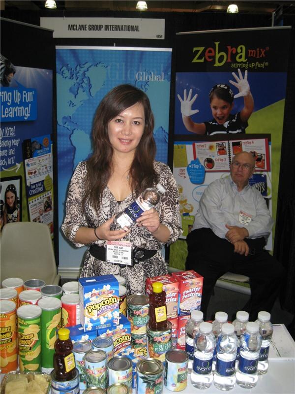 A woman holds an Evamor water bottle at the McLane Group International booth, with Zebra Mix banners behind her, a seated man at right, and Lady Liberty canned goods and Evamor bottles on the table.