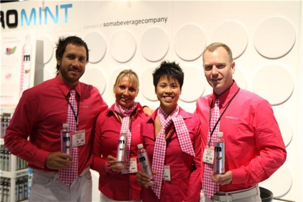 Four people in pink shirts and pink checked ties pose together, each holding a Metromint water bottle, in front of a white wall reading a product of somabeveragecompany.